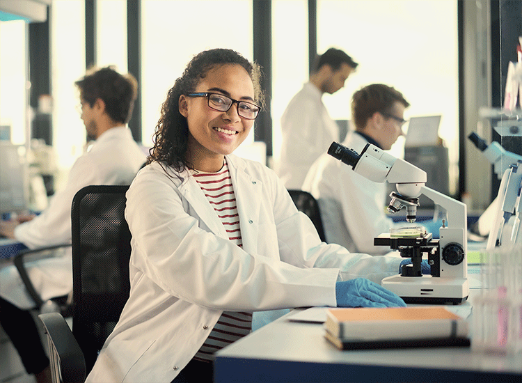 A woman works at a microscope in a biology classroom with other students.