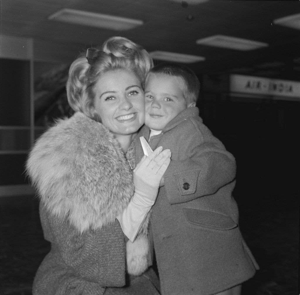 Jill Ireland with son Paul, both wearing woollen winter coats. Jill's coat has a large fur colour and she wears light gloves.