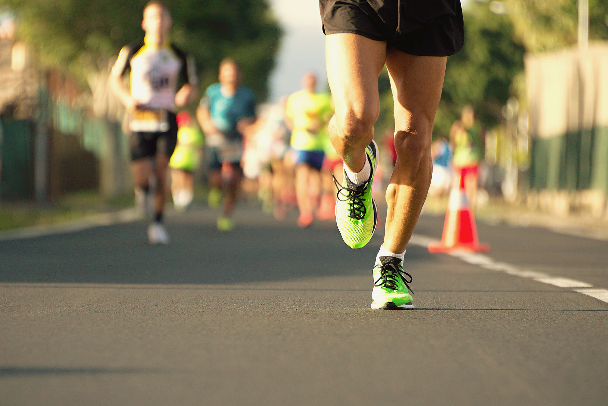 A group of people are seen running on a roadway. The focus of the image is on the runner in front, who is only visible from the waist down. They are wearing black shorts, white socks, and lime green sneakers.