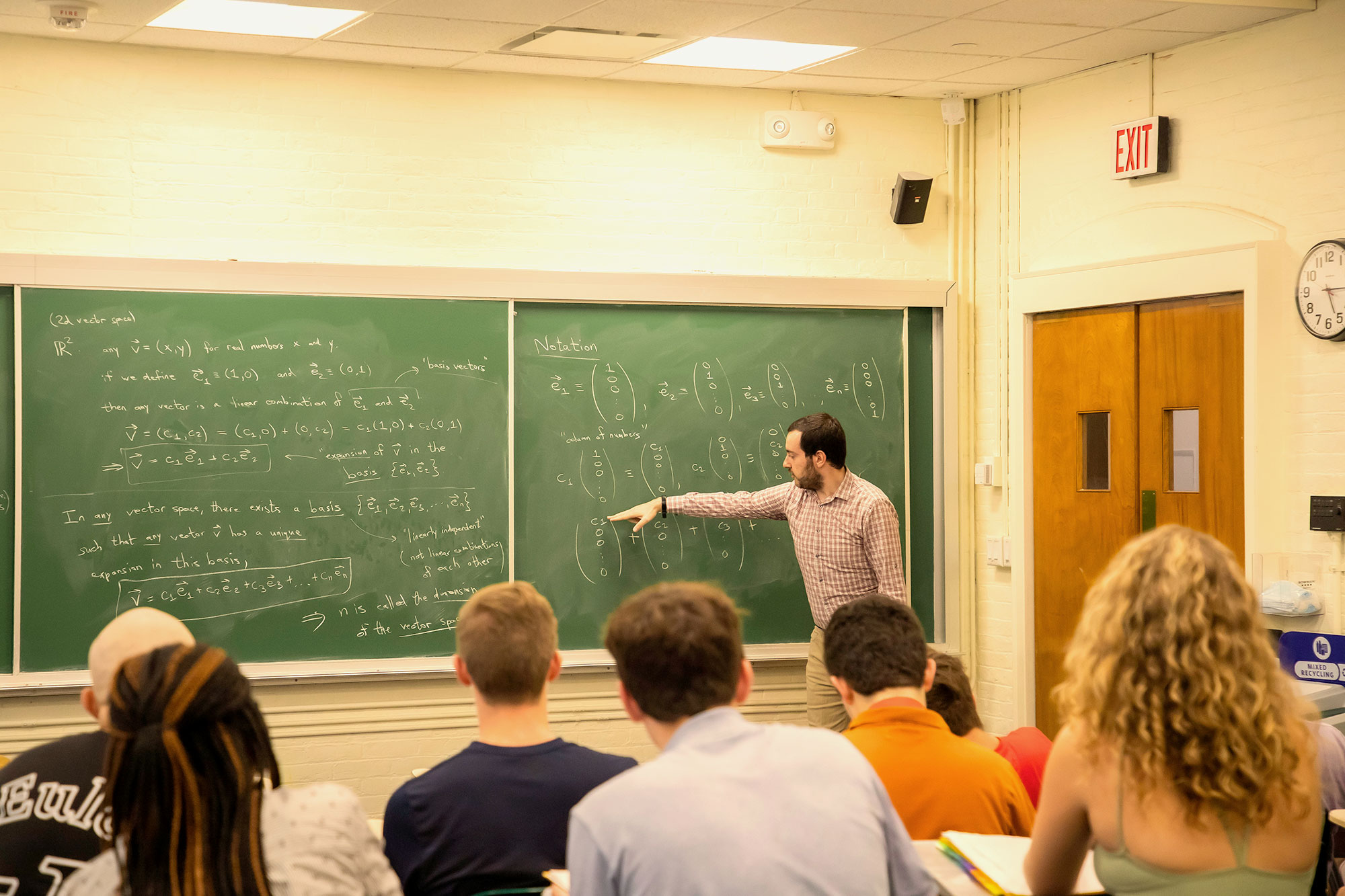 An instructor stands in front of a chalkboard with math symbols in front of a class of high school students.