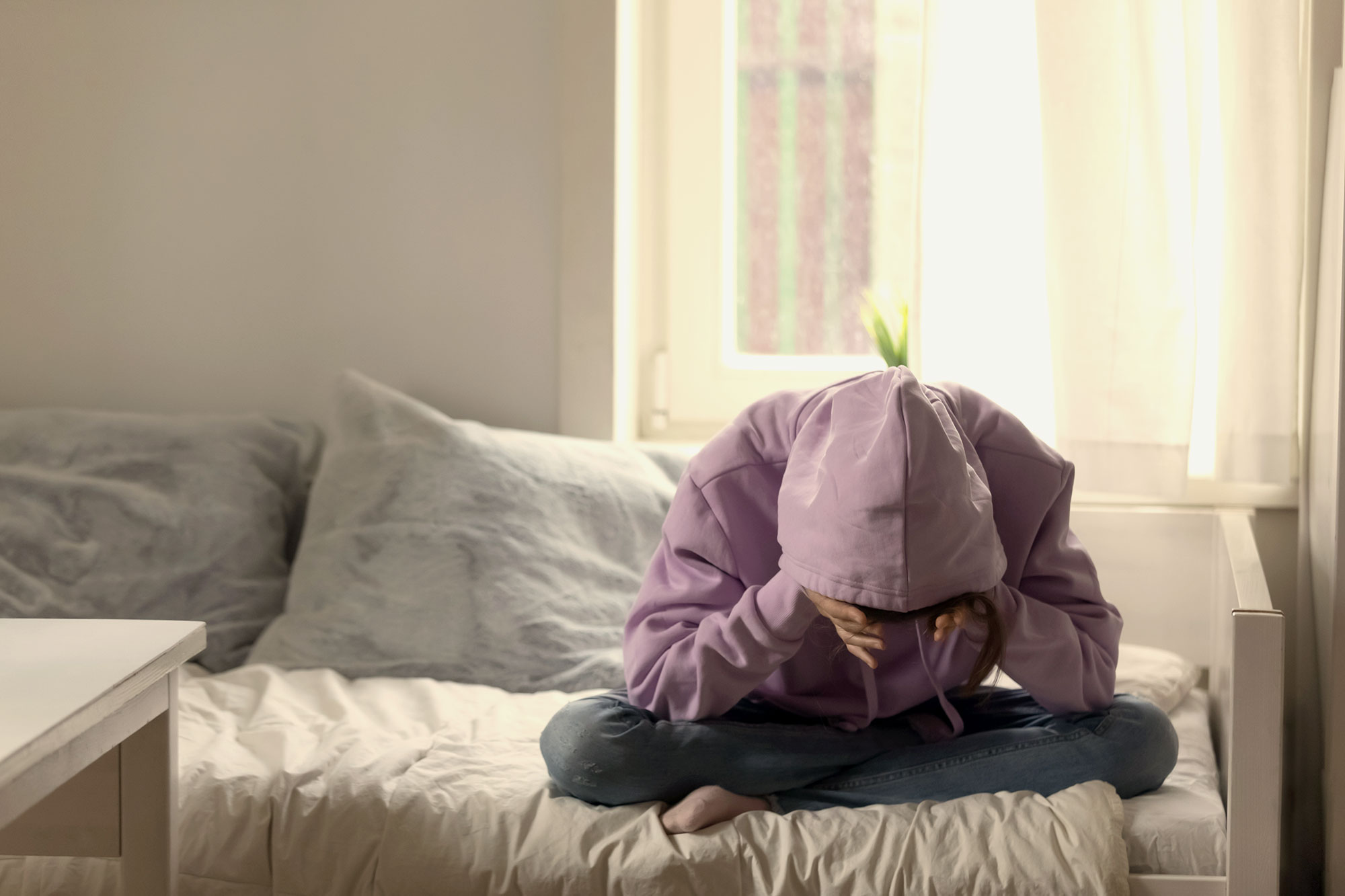 A students sits on a bed with their head in their hands, covered by a sweatshirt hood.