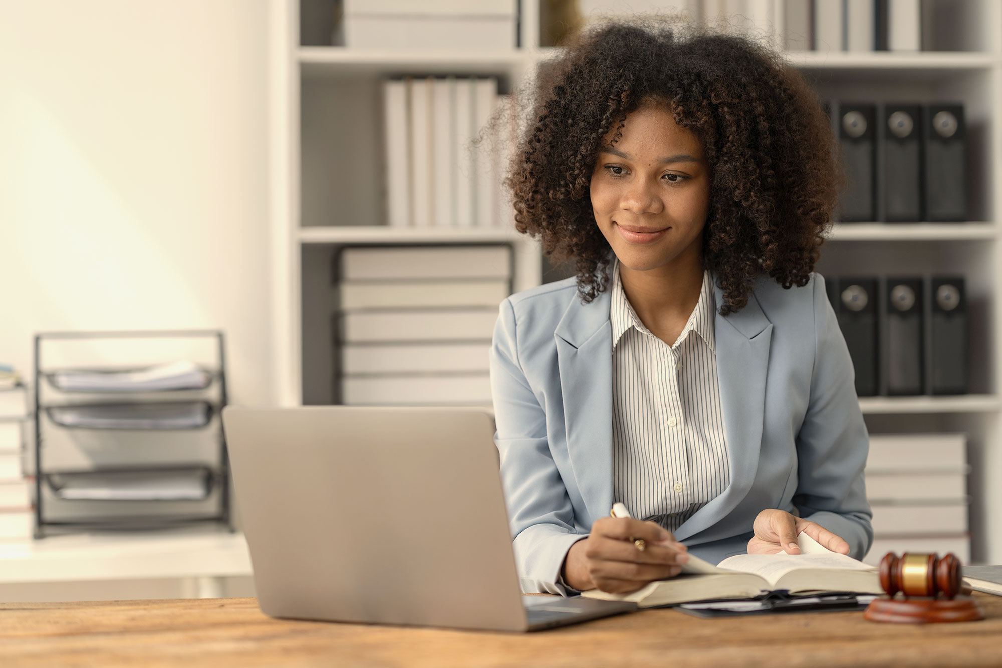 A smiling young woman works at a laptop while reading from a book.